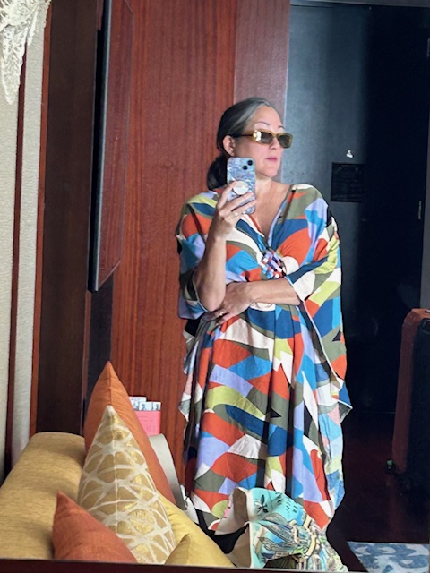 Woman in caftan standing with one arm crossed and other hand holding a cell phone.