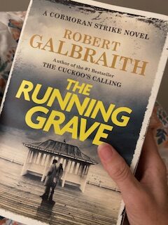 A hand holding Robert Galbraith's novel, The Running Grave. 